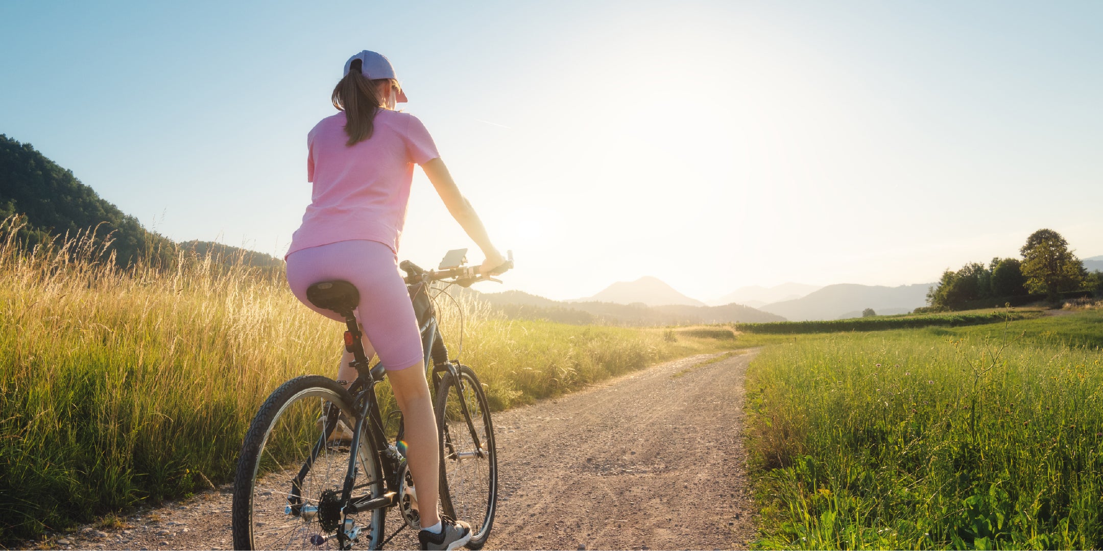 Femme en tenue de sport rose faisant du vélo sur un chemin de campagne au coucher du soleil, illustrant la reprise d’activité physique grâce à un soulagement articulaire au genou.