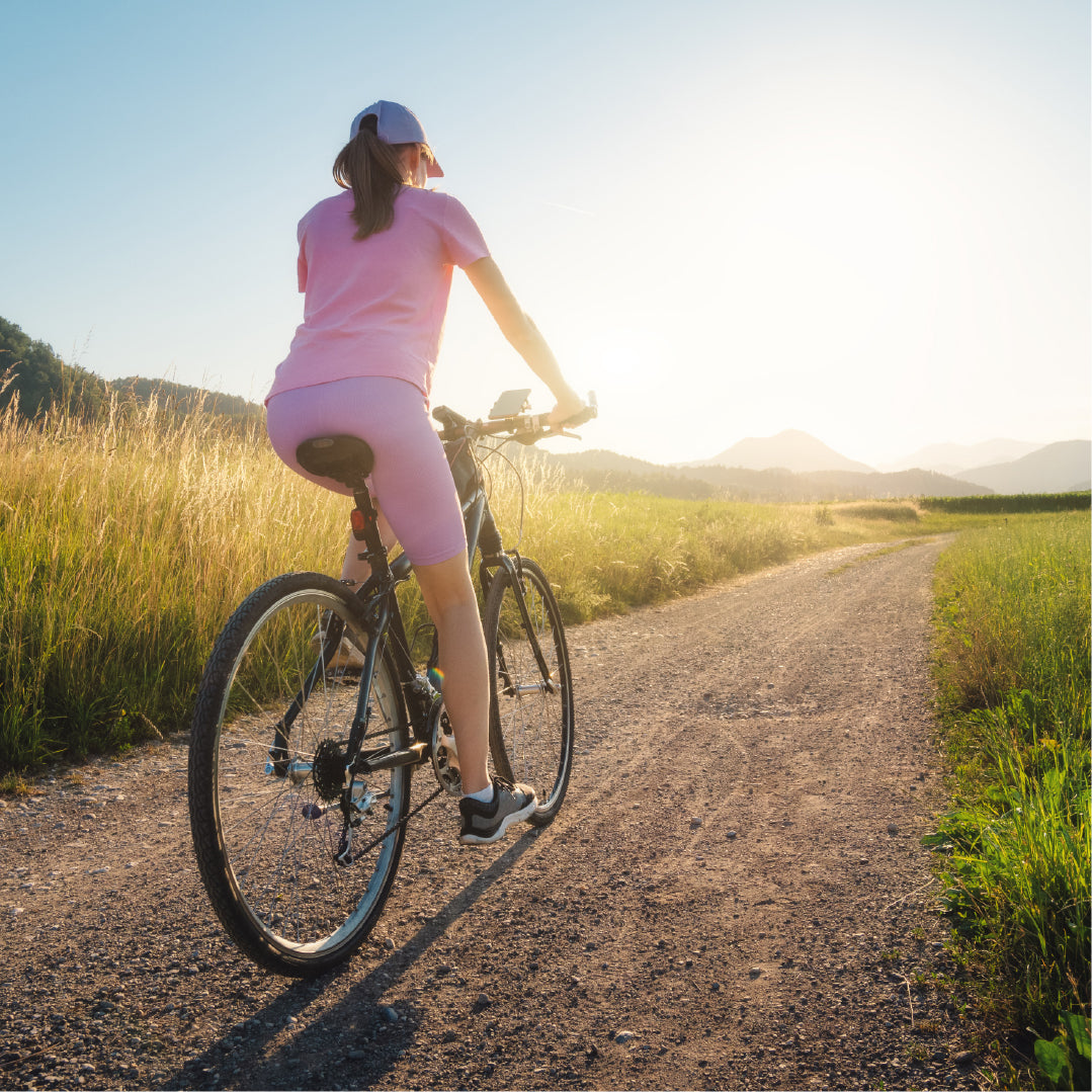 Femme en tenue de sport rose, pédalant à vélo sur un chemin de campagne au coucher du soleil, évoquant la mobilité retrouvée.